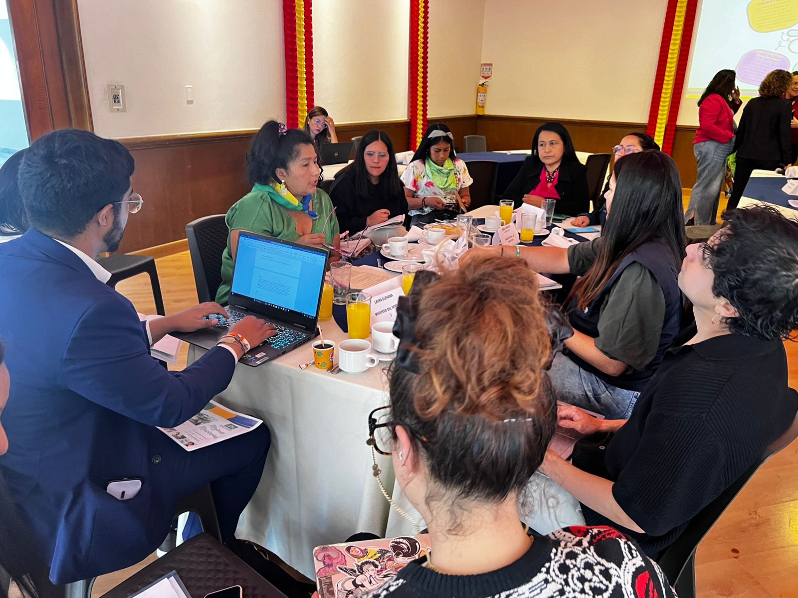 La fotografía muestra a un grupo de mujeres sentadas alrededor de una mesa y un hombre tomando notas en su portatil. Una mujer indígena vestida de verde habla y el resto escucha su intervención.